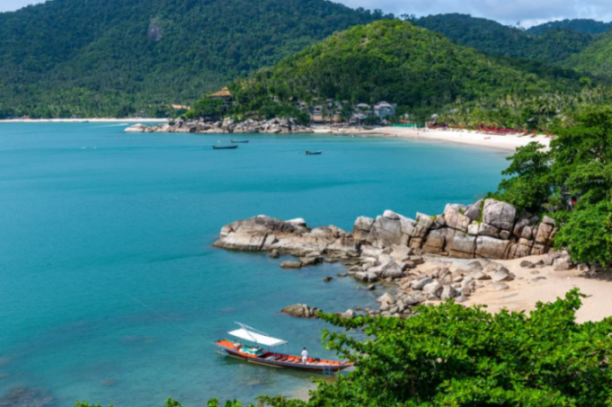A long-tail boat anchored in turquoise waters near a rocky, sandy beach backed by green hills and beachfront buildings.