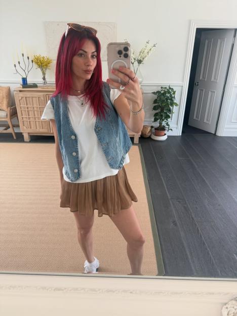 Selfie of a woman with red hair wearing a denim vest and brown shorts.
