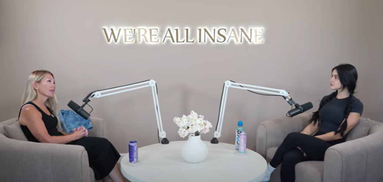 Two women sitting in chairs at a table with microphones, under a sign that reads "WE'RE ALL INSANE".