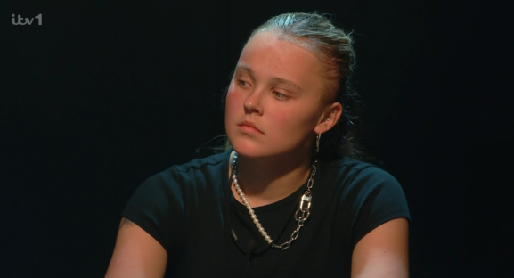 Screenshot of a young woman with light skin and dark hair wearing a black t-shirt and necklaces, looking thoughtfully to her left.