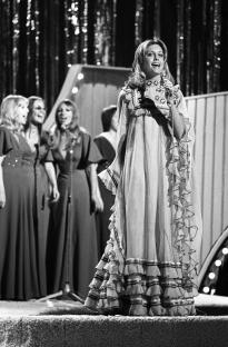 Black and white photo of Olivia Newton-John performing on stage in a ruffled dress.