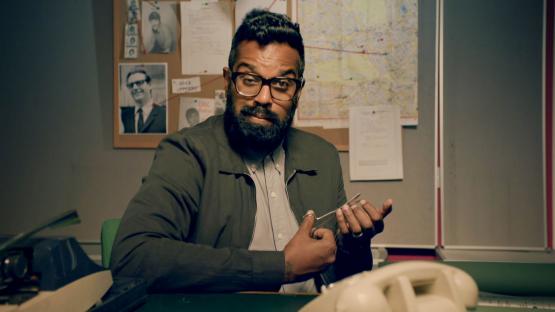 Romesh Ranganathan holding scissors in a detective's office.