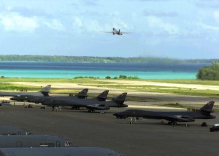 Photo of B-1 bombers on a runway with a B-1 taking off in the background, overlooking a tropical atoll.