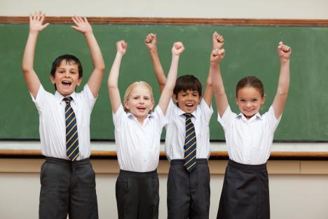 Four children in school uniforms with their arms raised in celebration in front of a chalkboard.