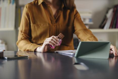 Businesswoman eating a protein bar while working on a tablet.