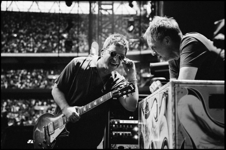 Black and white photo of two men at a concert; one plays guitar, the other looks on.