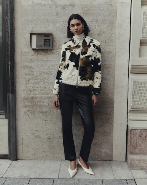 A woman modeling the M&S Autumn Winter 2025 collection wearing a white turtleneck, a faux fur jacket with a black, white, and brown cow print, black leather pants, and white pointed-toe heels.