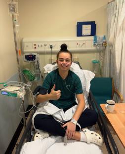A young woman gives a thumbs-up while sitting in a hospital bed.