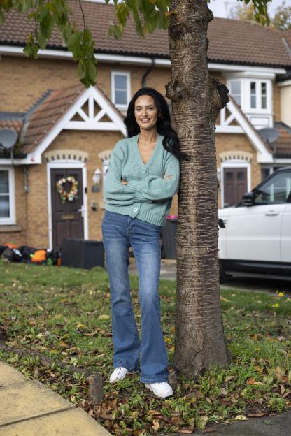 A woman in a blue cardigan and jeans leaning against a tree.