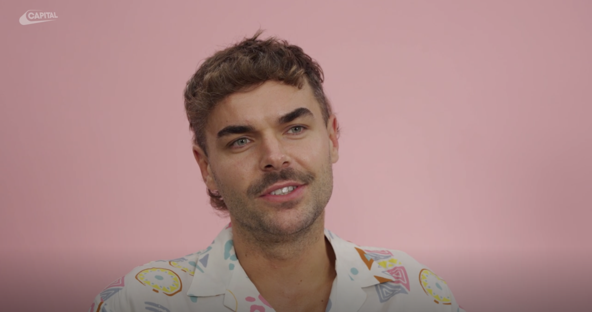 Man with styled hair and mustache, wearing a patterned shirt, against a pink background.