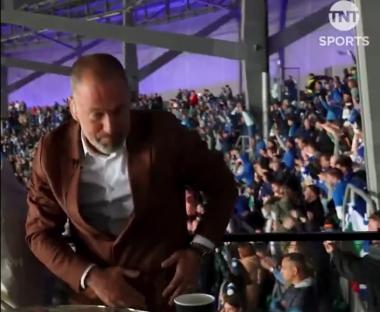 Man in brown jacket sits before a cheering crowd.