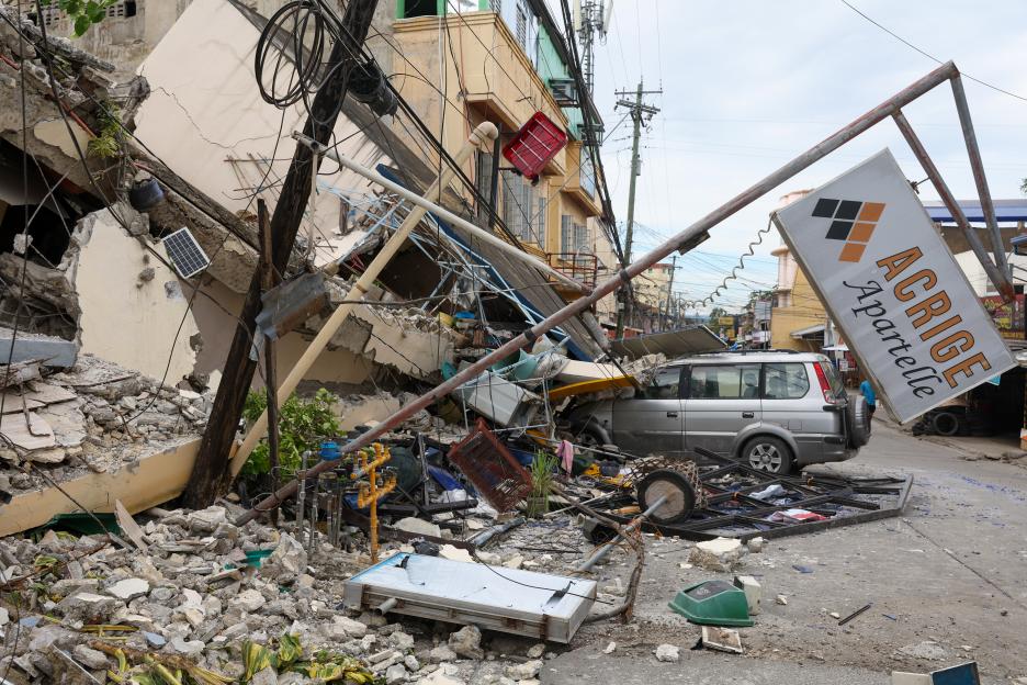 A gray car crushed by the debris of a partially collapsed building.