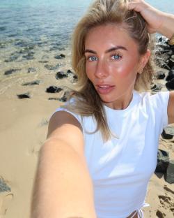 Selfie of a blonde woman on a beach wearing a white shirt.
