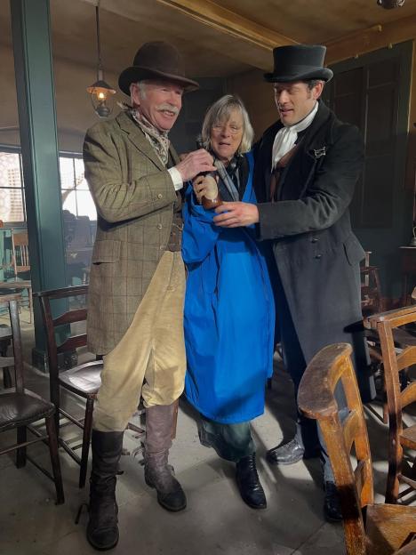 Hugh Norton, an older man with a mustache, and James Norton, a younger man, both in period costume, support an older woman in a blue coat and glasses.