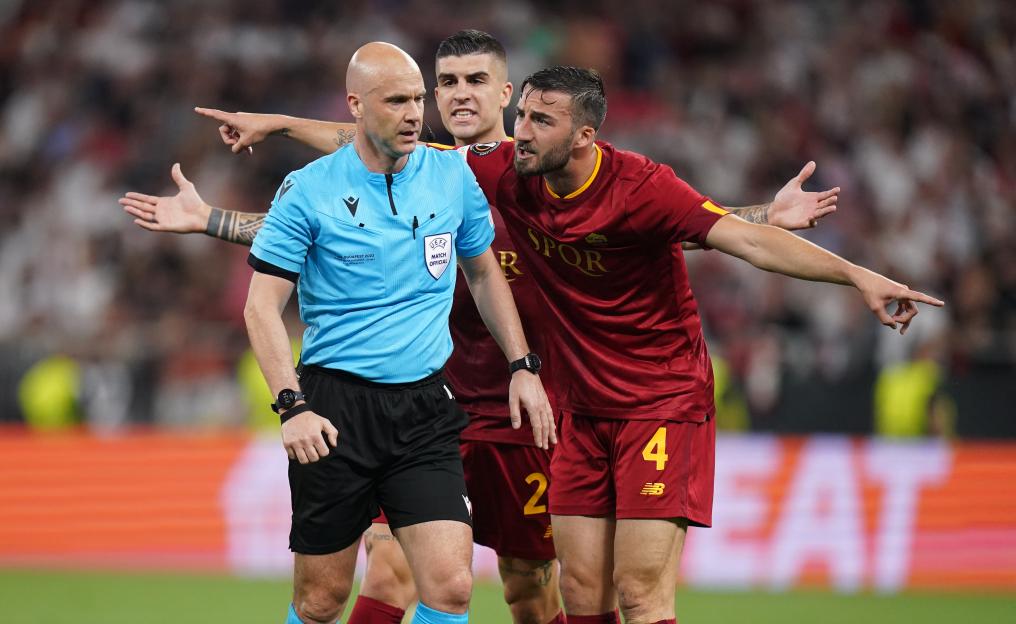 Referee Anthony Taylor surrounded by Roma players who are arguing before he reviews a penalty decision.