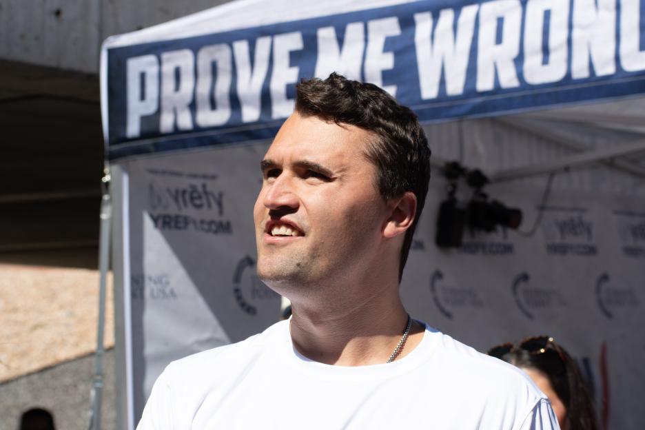 Charlie Kirk looking upward with a "Prove Me Wrong" banner in the background.