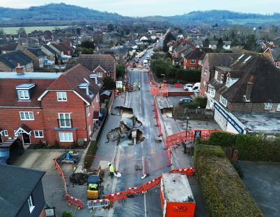 Aerial view of a large sinkhole in a residential street, closed off with barriers and emergency personnel on site.