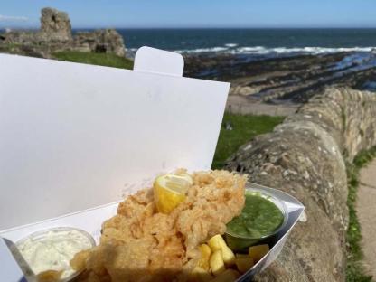 Fish and chips with tartar sauce and mushy peas, eaten outdoors near a stone wall and ocean.