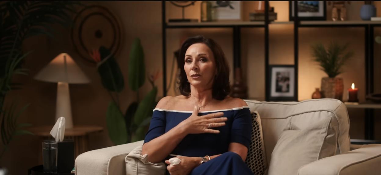 A woman with dark hair in a blue dress sits on a couch, holding her chest with one hand.