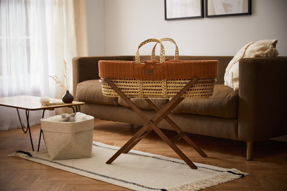 Wicker baby bassinet with terracotta knit liner on wooden stand.