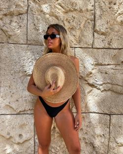 Woman in a black bikini holding a large straw hat in front of a stone wall.