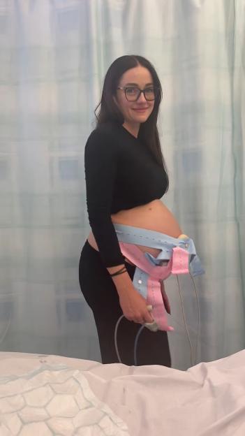 Pregnant woman with monitor straps around her belly, smiling in a hospital room.