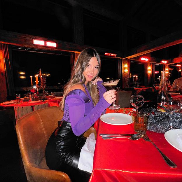 Laura Villars wearing a purple top and black leather skirt, holding a cocktail at a restaurant with red tablecloths.