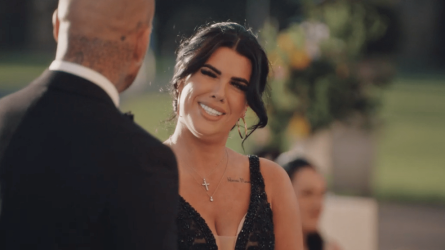 A smiling woman in a black sequined dress and a cross necklace looks at a man in a tuxedo.