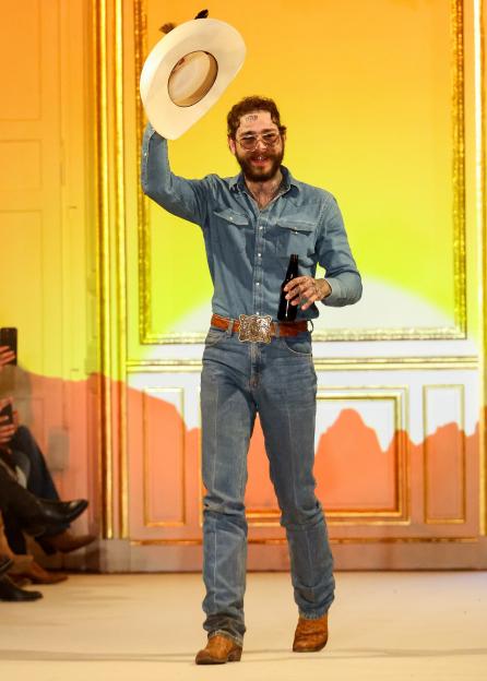 Post Malone walking a runway, holding a beer bottle and raising his cowboy hat.