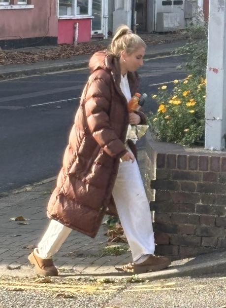 Stacey Solomon in a long brown puffer coat, white pants, and brown shoes, carrying a drink and a bag, walking on a street next to a brick wall.