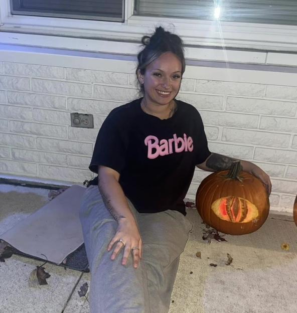 A young woman wearing a Barbie t-shirt sits next to a carved pumpkin with glowing eyes.