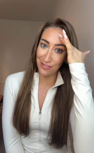 A woman with long brown hair, wearing a white athletic top, touches her forehead.