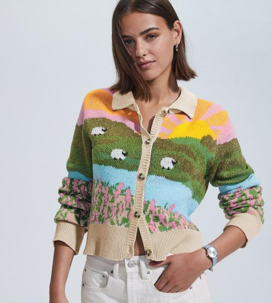 Woman in a knit cardigan with a pastoral landscape scene and white jeans.