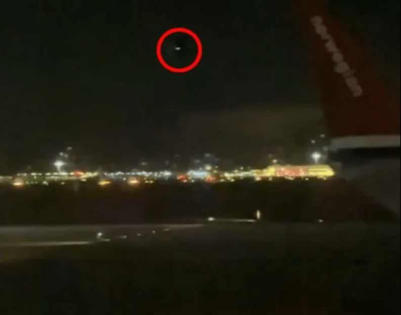 Nighttime image of an object in the sky circled in red, above a cityscape with a plane wing in the foreground.