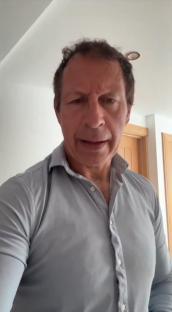 A man in a light gray shirt speaks to the camera.
