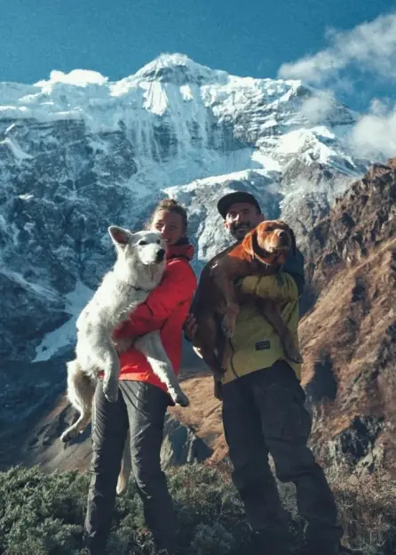 Two hikers holding dogs in front of a snowy mountain.
