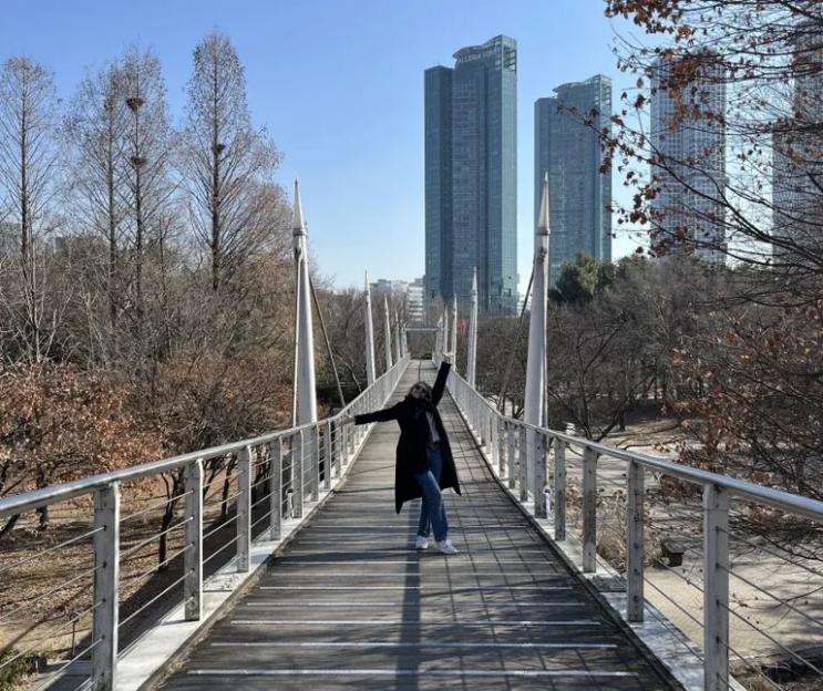 A person with outstretched arms stands on a long pedestrian bridge with tall buildings in the background.
