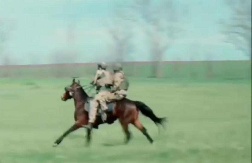 Two soldiers in military fatigues and helmets on horseback gallop across a green field.