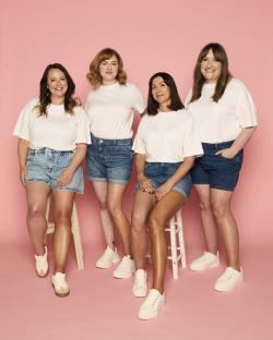 Four women in white t-shirts and denim shorts pose for a photo.