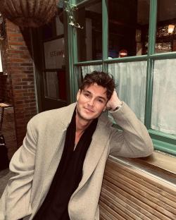 Man in a tan coat sitting on a bench outside a building.
