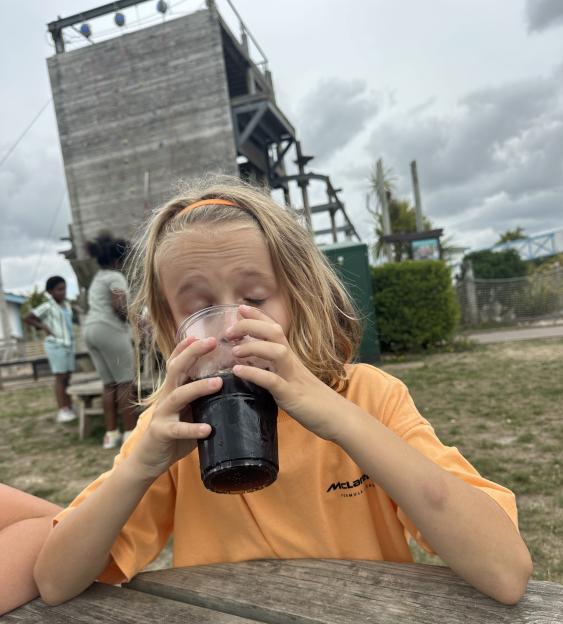 Child drinking from a cup outdoors near a climbing wall.