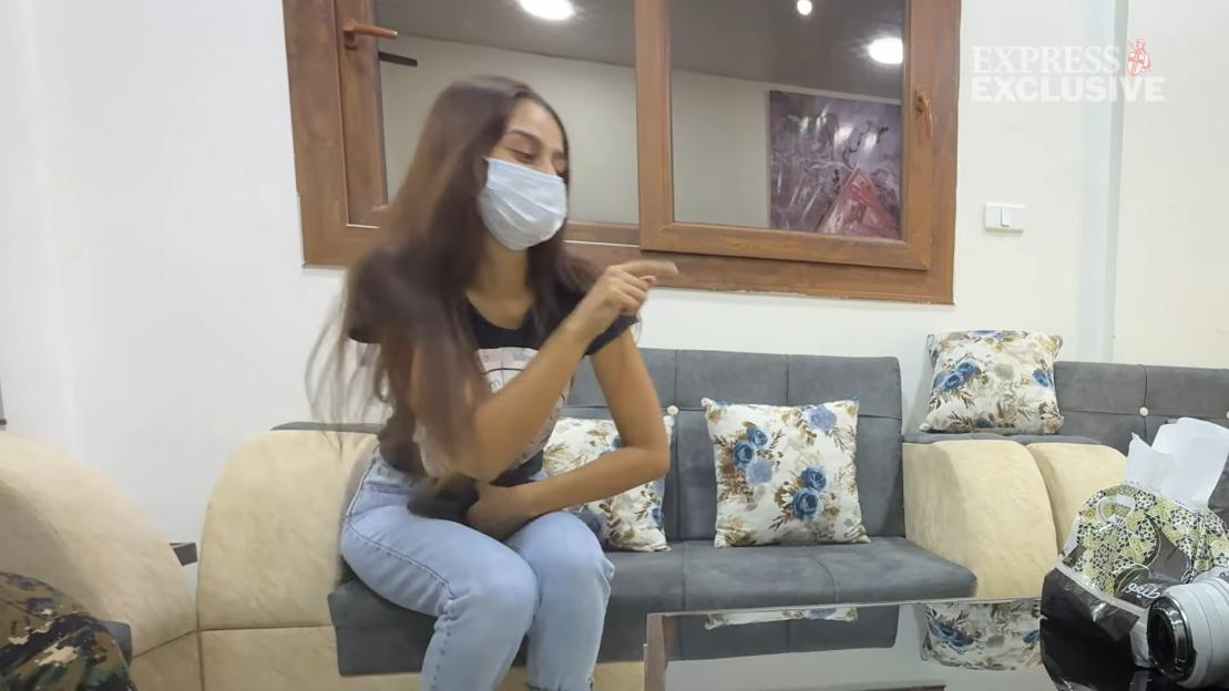 Shamima Begum wearing a face mask, seated on a couch, pointing and looking away.