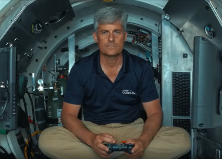 Man in OceanGate shirt sitting in a submersible, holding a controller.