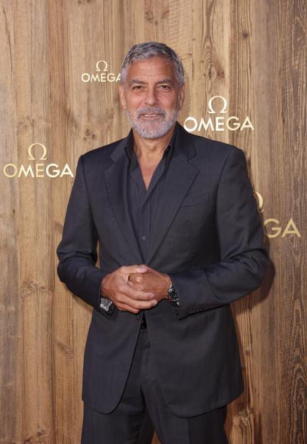 George Clooney in a dark suit against a wooden wall with Omega logos.