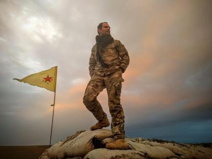 A soldier in camouflage stands on a sandbag barricade with a yellow flag in the background.
