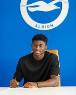A young man signing a document at a table, with the Brighton & Hove Albion logo in the background.
