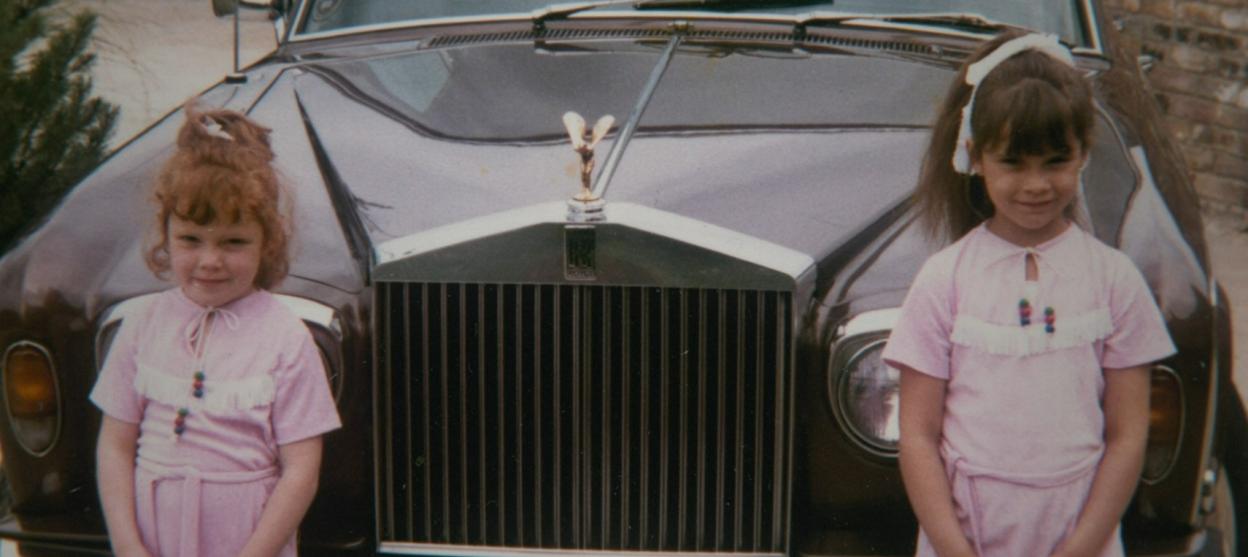 Two young girls in pink dresses standing in front of a dark luxury car.