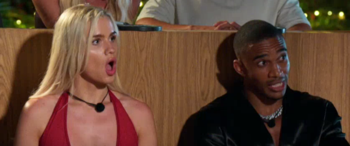 A woman with blonde hair in a red dress with a surprised expression next to a man with dark skin and hair in a black shirt.