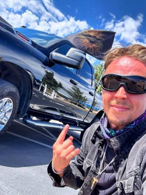 Man in sunglasses making a hand gesture in front of a black Ram truck.