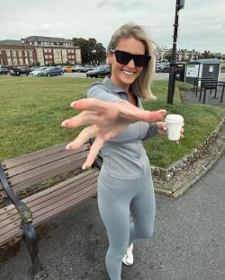 Woman in gray athleisure outfit reaching toward the camera, holding a coffee cup.